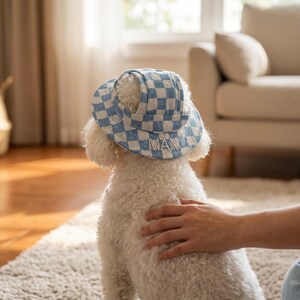 以下が含まれることがあります： 青と白のチェック柄の帽子をかぶった白い犬。つばには「MAX」という名前が刺繍されています。犬は人に撫でられています。この帽子はペット用にデザインされています。