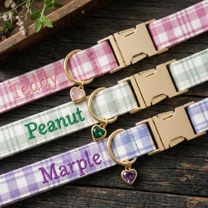 May include: Three plaid dog collars in pink, green, and purple, each with a gold-colored buckle and a heart-shaped charm. The collars are embroidered with the names "Teddy", "Peanut", and "Marple".