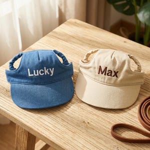 Può includere: Due cappellini da baseball per cani. Uno è blu denim con "Lucky" ricamato in bianco. L'altro è beige con "Max" ricamato in marrone. Entrambi hanno fori per le orecchie e una visiera curva. Sono visibili anche una pallina da tennis e un guinzaglio in pelle.