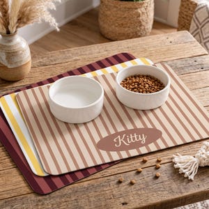 May include: Three rectangular pet placemats with rounded corners, featuring vertical stripes in maroon, yellow, and tan. The top tan mat displays an oval patch with the word Kitty in white script. Two white ceramic bowls sit on the top mat.