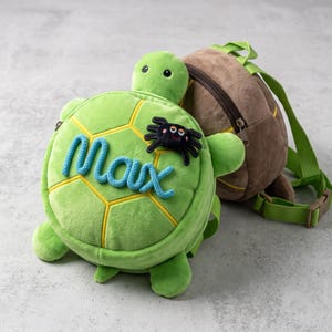 May include: A plush turtle-shaped backpack featuring a bright green shell with yellow piping and the name Max embroidered in blue script. A small black spider applique with white eyes is attached to the shell. The back section is brown plush.