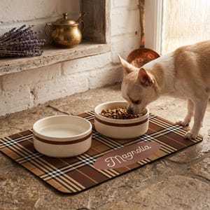 May include: A rectangular pet feeding mat featuring a brown, black, and white plaid pattern with a tan oval accent containing the name Magnolia in white script. Two round, cream-colored ceramic bowls sit on the mat, one filled with dry food.