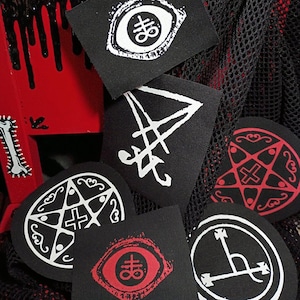 May include: A collection of black patches with white and red occult symbols. The patches feature various designs, including an eye, a pentagram, and other geometric shapes. The background includes a red and black decorative element and a black mesh.