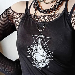 Könnte beinhalten: Ein schwarzes Tanktop mit einem weißen Grafikdruck einer Hand, umgeben von geometrischen Formen und einem Augensymbol. Das Oberteil hat einen Metallring am Ausschnitt und wird über einem schwarzen Langarmshirt aus Netzstoff getragen. Dazu werden eine mehrlagige schwarze Kettenhalskette und ein schwarzes Halsband getragen.