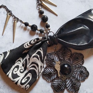 May include: A necklace featuring a black chain with silver-toned spikes and black textured beads. A black fabric bow with a white abstract pattern is attached to a large, dark, filigree flower pendant with a central black faceted stone.