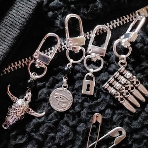May include: A collection of silver-toned keychains with various charms, including a bull skull, an eye symbol, a padlock, and a cluster of skulls. The keychains are displayed on a black, textured surface with a zipper and safety pins.