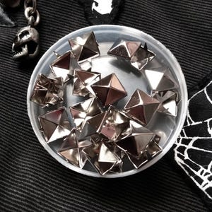 May include: A collection of metallic silver pyramid-shaped studs contained within a small, clear, circular plastic container. The studs feature a four-sided pointed top and a reflective, polished finish, arranged in a cluster inside the container.
