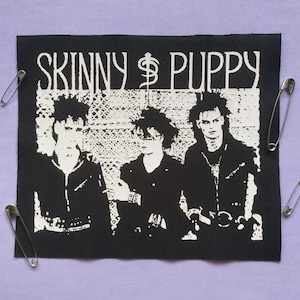 Op de afbeelding: Zwart-wit patch met de band Skinny Puppy met drie figuren. De naam van de band staat boven de figuren gedrukt. Zilveren veiligheidsspelden omringen de patch op een lavendelkleurige achtergrond.