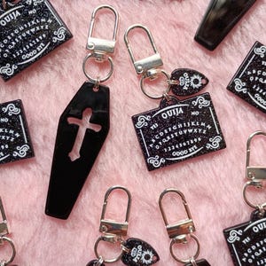 Op de afbeelding: Zwarte Ouija bord en doodskistvormige sleutelhangers met zilverkleurige sluitingen. De sleutelhangers tonen de tekst "OUIJA" en het alfabet, cijfers en de woorden "YES", "NO" en "GOOD BYE". De doodskist sleutelhangers hebben een kruis uitsparing.