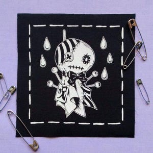 Könnte beinhalten: Ein schwarzer quadratischer Aufnäher mit einer weißen Cartoon-Voodoo-Puppe-Illustration, umgeben von Tränen und einem weißen Nahtrand. Sicherheitsnadeln sind auf einem violetten Hintergrund verstreut.