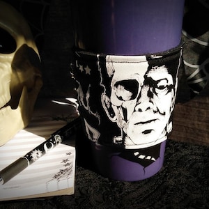 May include: A black and white fabric coffee cup sleeve with a graphic of Frankenstein's monster. The sleeve is on a purple coffee mug.