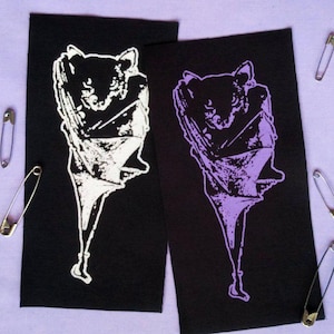 May include: Two black fabric patches featuring a bat design, one in white and the other in purple, are shown on a light purple surface. Several safety pins are scattered around the patches, adding a decorative element.