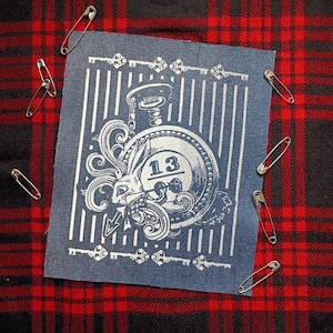 May include: A blue patch featuring a pocket watch design with a rabbit and the number 13. The patch has a striped background and key designs. Safety pins are scattered around the patch on a red and black plaid background.
