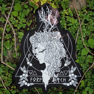 May include: A black patch with a white line art illustration of a profile of a figure wearing a floral crown and antlers. The design includes geometric triangles, botanical elements, and the text "FARM WITCH" at the bottom in a stylized font.
