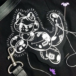 May include: A black fabric patch featuring a white graphic of a stitched-together creature wearing a muzzle. The patch is surrounded by three purple bat-shaped pins, silver sewing needles with white thread, and a black strap with a silver metal clip.