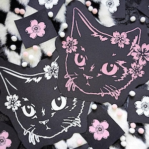 May include: Black cat-shaped patches with pink and white floral accents. The cat faces have floral crowns and are surrounded by small square patches with flower designs. White and pink pom-poms are scattered around the patches.