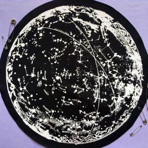 Może przedstawiać: Czarna i biała okrągła naszywka z mapą nieba, przedstawiająca konstelacje i wzory gwiazd. Naszywka jest otoczona czarną obwódką i ma kilka srebrnych agrafek. Tematem jest astronomia.