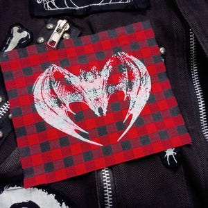 Op de afbeelding: Een rood-zwart geruit patch met een wit vleermuisontwerp met uitgespreide vleugels. De patch is genaaid op een zwarte jas met zilveren ritsen en andere decoratieve patches. Het thema is gothic of alternatieve mode.