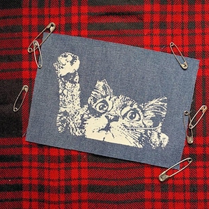 May include: A navy blue fabric patch with a white cat illustration, one paw raised. The cat's face is detailed. Silver safety pins surround the patch, set against a red and black plaid background. The patch is approximately 7 cm x 10 cm.