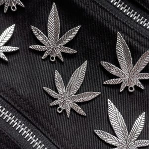 May include: A collection of silver-toned metal charms shaped like cannabis leaves, each featuring detailed vein textures and a small circular loop at the base for attachment, arranged on a black fabric surface with a visible diagonal zipper.