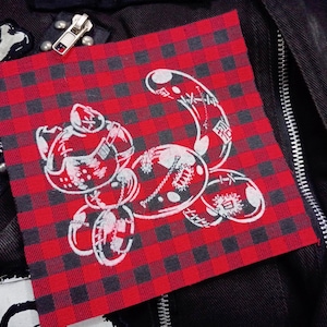 Peut inclure: Un écusson à carreaux rouges et noirs avec un dessin de chat de dessin animé blanc. Le chat est stylisé avec des formes arrondies et des détails internes. L'écusson est cousu sur un tissu noir, avec une fermeture éclair et des accents métalliques visibles.