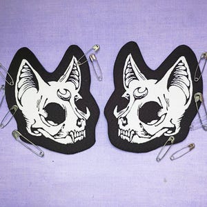 Może przedstawiać: Dwie czarne naszywki z czaszkami kotów z białymi detalami, każda ozdobiona agrafkami, spoczywają na lawendowej powierzchni. Naszywki przedstawiają szczegółowe ilustracje czaszek ze spiczastymi uszami i otwartymi ustami.