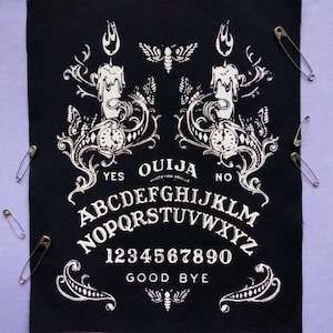 May include: Black Ouija board with white lettering and decorative elements, including candles, moths, and ornate designs. The board features the alphabet, numbers, and the words "Yes," "No," and "Good Bye." Safety pins are scattered around the board.