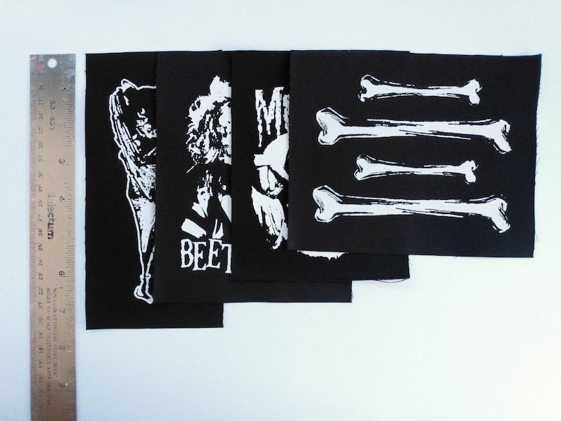 May include: Black fabric patches with white graphic designs. The patches feature illustrations of figures, the word "BEET", and bone shapes. A metal ruler with inch measurements is on the left.