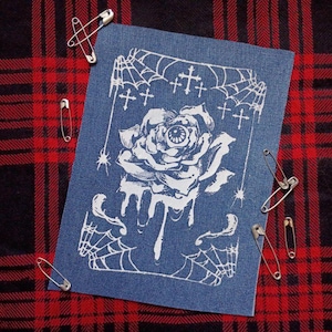 Peut inclure: Un écusson en jean bleu avec un motif blanc d'une rose avec un œil, des toiles d'araignées et des croix. Des épingles de sûreté argentées sont disposées autour de l'écusson sur un fond à carreaux rouge et noir.