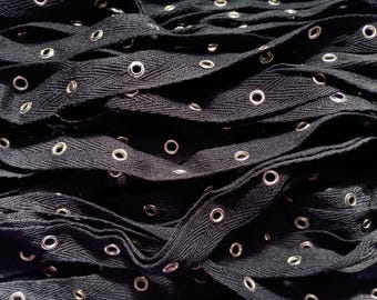 Puede incluir: Primer plano de cordones negros con ojales plateados. Los cordones están tejidos en un patrón de espiga y están superpuestos, creando un efecto texturizado. Los ojales están espaciados uniformemente y añaden un contraste metálico a la tela oscura.