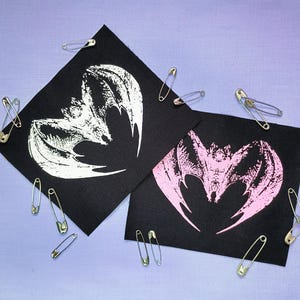 May include: Two black squares with bat designs in white and pink, surrounded by safety pins. The bat images have detailed wing patterns. The background is a light purple fabric.