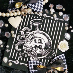 Pode incluir: Um cartão em preto e branco com o tema Alice no País das Maravilhas com um design de relógio de bolso com um coelho e o número 13. O cartão é decorado com pérolas, pedras preciosas, chaves e um coração branco. Também estão presentes dois laços quadriculados e uma rosa branca.