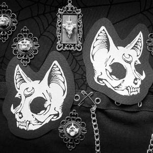 May include: Black and white image featuring gothic-themed accessories. Two patches with cat skull designs are displayed alongside ornate brooches and a framed pendant. The items are arranged on a black fabric with a spiderweb pattern.