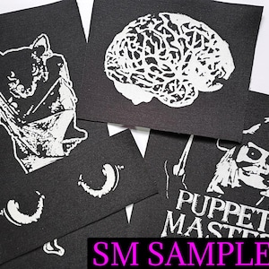 May include: A collection of black fabric patches with white graphic designs. The patches feature a bat, a brain, a character from "Puppet Master", and other abstract shapes. The text "SM SAMPLE #2" is visible in the image.
