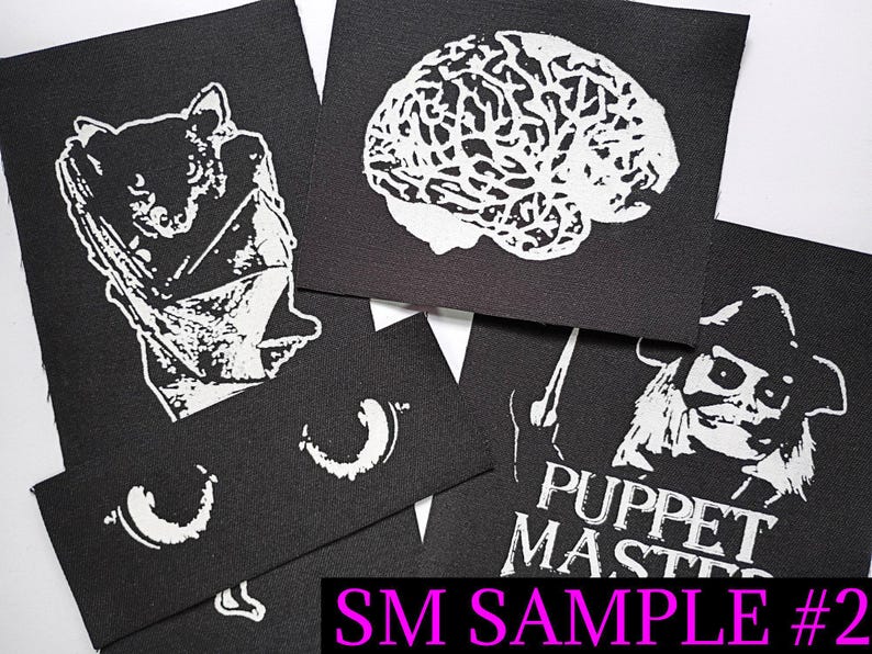 May include: A collection of black fabric patches with white graphic designs. The patches feature a bat, a brain, a character from "Puppet Master", and other abstract shapes. The text "SM SAMPLE #2" is visible in the image.