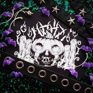 May include: A black and white Halloween-themed illustration featuring a pumpkin with a stitched mask, candles, and a tombstone. The image is surrounded by purple bat-shaped decorations, bone accents, and a black belt with silver grommets and star studs.