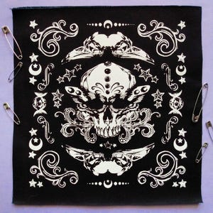May include: Black square fabric featuring a white skull design, stars, moons, and decorative swirls. Several silver safety pins are placed around the edges. The design has a gothic or occult theme.