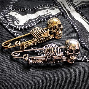 May include: Two skull-shaped safety pins. One is gold, the other silver. Both pins have a skeleton design. They rest on black fabric with white stitching and a decorative patch.