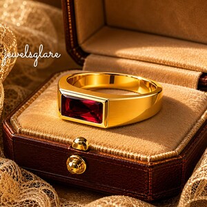 Könnte beinhalten: Ein goldener Siegelring mit einem rechteckigen, tiefroten Edelstein, der horizontal in der Mitte gefasst ist. Der Ring hat eine polierte, glatte Oberfläche und liegt in einer braunen, samtgefütterten Schatulle mit dem Text jewelsglare.
