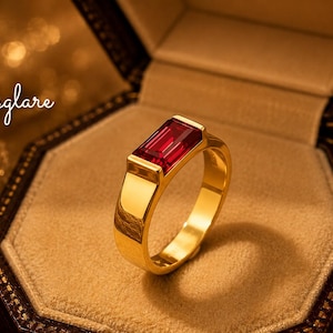 May include: A gold ring featuring a rectangular, emerald-cut red gemstone set in a bezel setting. The ring is displayed inside an open, dark-colored, octagonal jewelry box with a tan interior. The text "jewelsglare" is visible in the upper left corner.