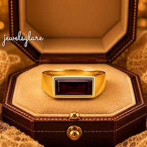 May include: A gold ring featuring a horizontal rectangular deep red gemstone set in the center. The band is smooth and polished, showcasing a clean design. The text "jewelsglare" is visible in the upper left corner of the image.