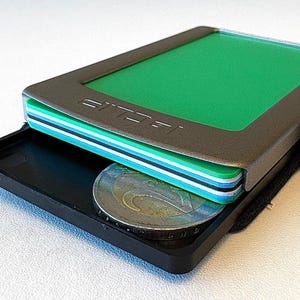 Könnte beinhalten: Ein metallgraues Portemonnaie mit grünem Innenraum und einem Stapel farbiger Karten. Das Portemonnaie ist geöffnet und zeigt eine schwarze Ablage mit einer Münze. Das Wort "JELP" ist sichtbar.