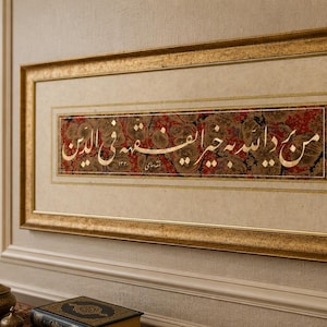 Puede incluir: Una obra de arte enmarcada con caligrafía árabe. El marco dorado rodea un panel central con escritura blanca y dorada sobre un fondo estampado en rojo, marrón y crema. La obra está colgada en una pared beige.