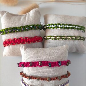 Peut inclure: Plusieurs bracelets macramé faits à la main de différentes couleurs, dont vert, rouge et rose, sont présentés sur de petits coussins carrés blanc cassé. Les bracelets présentent des nœuds complexes et sont ornés de perles. L'arrière-plan est de couleur neutre.