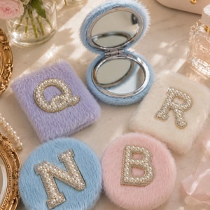 Op de afbeelding: Een verzameling zachte, pastelkleurige compacte spiegels en etuis. De open blauwe compacte spiegel heeft een zilveren frame. De etuis zijn lavendel, wit, roze en blauw, elk versierd met een met parels bezette letter: Q, R, N en B. Een klein kaartje leest "Have a nice day."