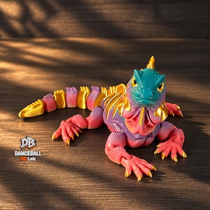 Op de afbeelding: Een gearticuleerd leguaanfiguur met een blauwgroene kop, een verlooplichaam van roze naar paars, en gouden accenten op de stekels, klauwen en rug. De tekst "DB DANCEBALL 3D Lab" staat in zwart en oranje gedrukt aan de linkeronderzijde.