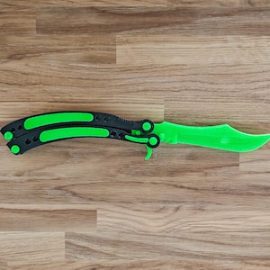 Könnte beinhalten: Ein Butterflymesser mit neongrüner Klinge und Akzenten sowie einem schwarzen Griff. Das Messer ist geöffnet und zeigt die volle Länge von Klinge und Griff. Das Messer liegt auf einer Holzoberfläche.