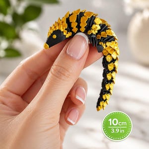 Könnte beinhalten: Eine kleine, bewegliche Pangolin-Figur mit schwarzer Grundfarbe und metallisch goldenen Akzenten auf den Schuppen. Die Figur ist C-förmig gebogen und enthält ein kreisförmiges grafisches Etikett, das eine Länge von 10 cm angibt.