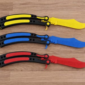 Puede incluir: Tres navajas mariposa con mangos negros y hojas amarillas, azules y rojas. Las navajas están sobre una superficie de madera. Los mangos tienen detalles de color que coinciden con el color de la hoja.