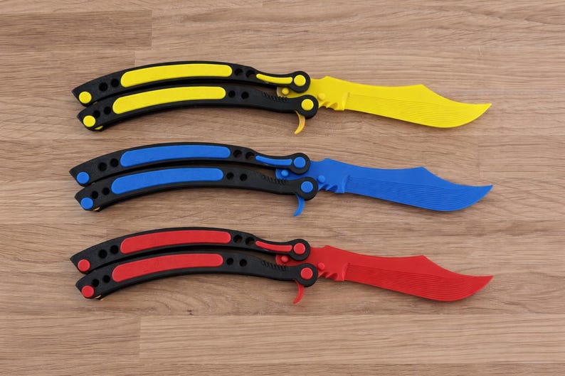 Op de afbeelding: Drie vlindermessen met zwarte handvatten en lemmeten in geel, blauw en rood. De messen liggen op een houten oppervlak. De handvatten hebben gekleurde accenten die passen bij de kleur van het lemmet.